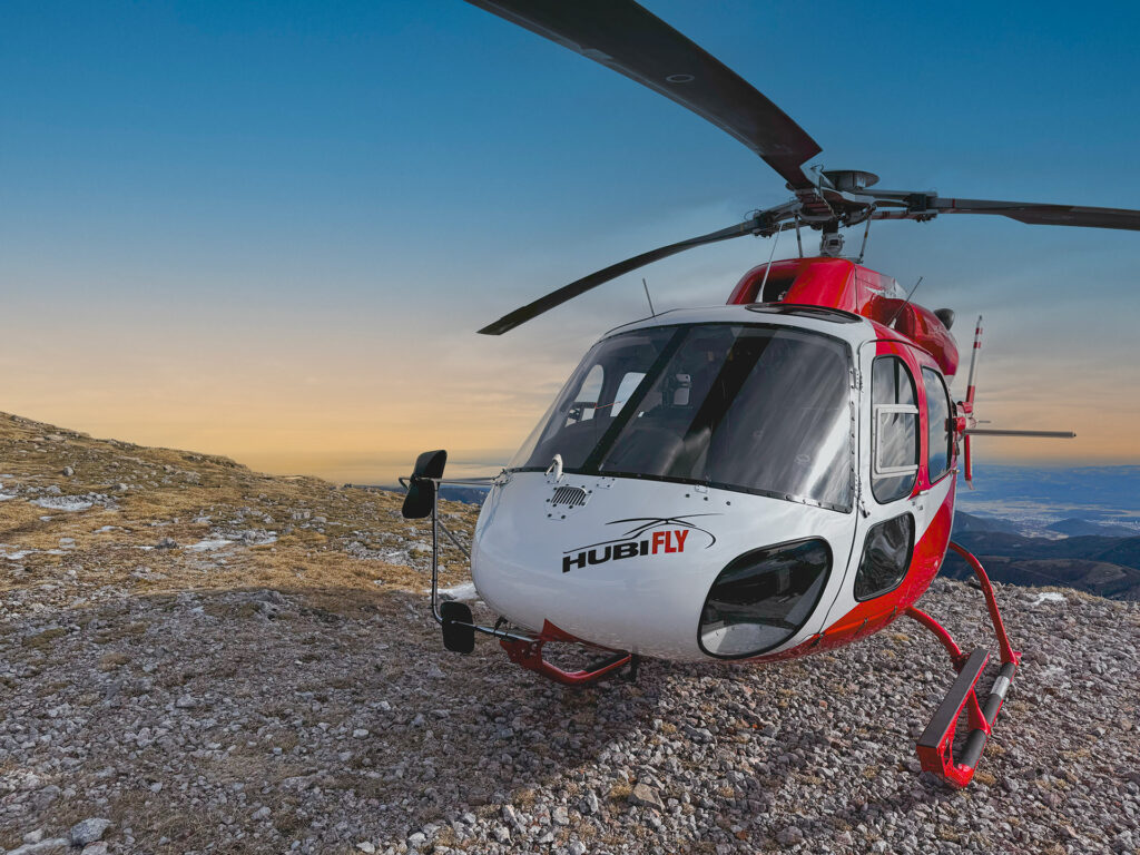 Rot weiß roter Hubschrauber vom Type AS350 mit HubiFly Logo, abgestellt auf einem Bergkamp. Im Hintergrund Sonnenuntergangsstimmung.