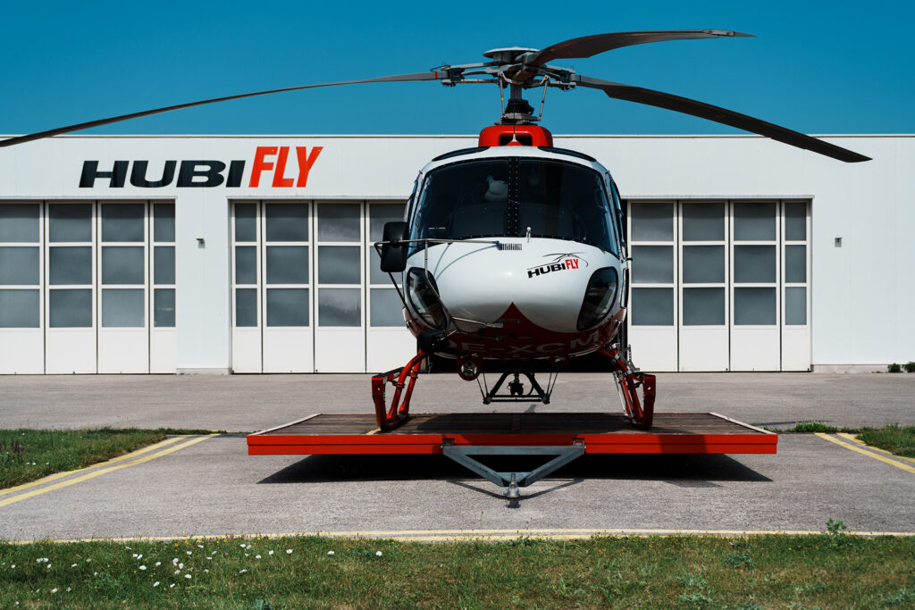 Foto eines AS350 B2 in rot weiß, im Hintergrund Hubifly Hangar und Gebäude