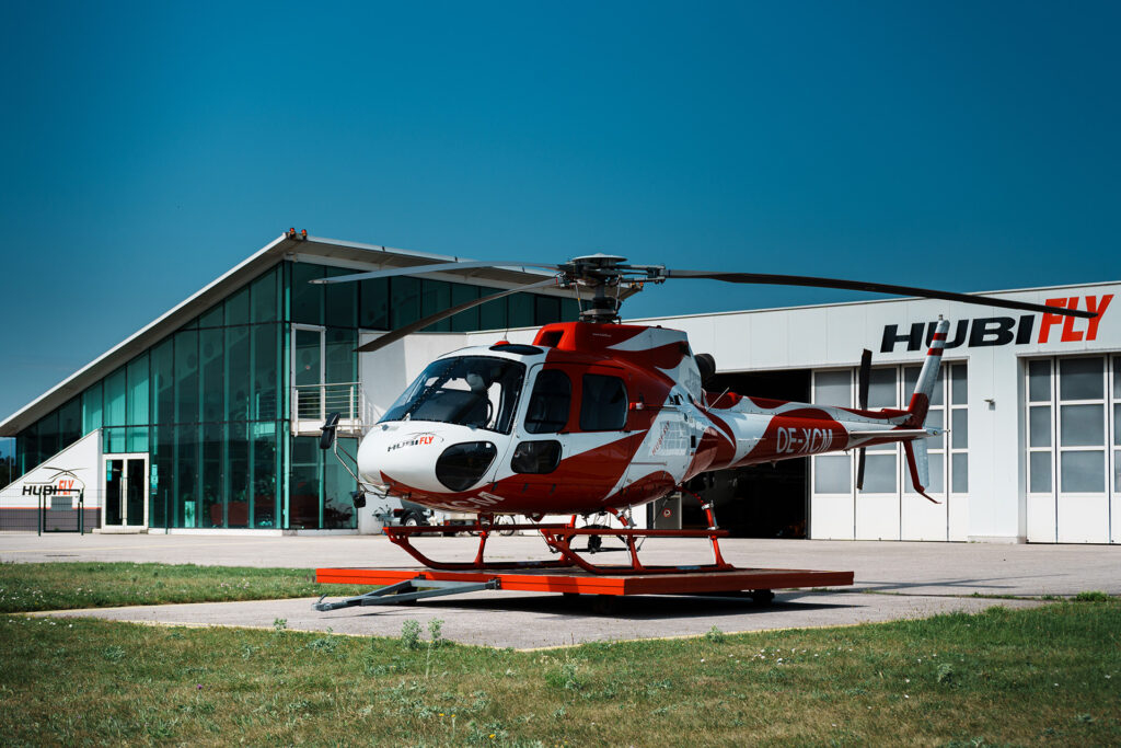 Schrägansicht eines AS350 B2 in rot weiß, im Hintergrund Hubifly Hangar und Gebäude