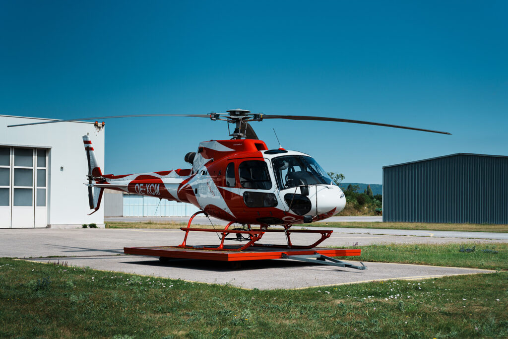 Schrägansicht eines AS350 B2 in rot weiß, im Hintergrund Hubifly Hangar und Gebäude