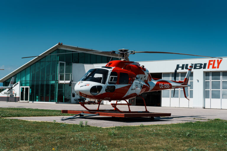 Schrägansicht eines AS355 F2 in rot weiß, im Hintergrund Hubifly Hangar und Gebäude