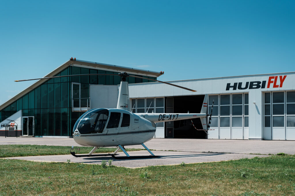 Schrägansicht eines Robinson R44 Raven II in weiß, im Hintergrund Hubifly Hangar und Gebäude