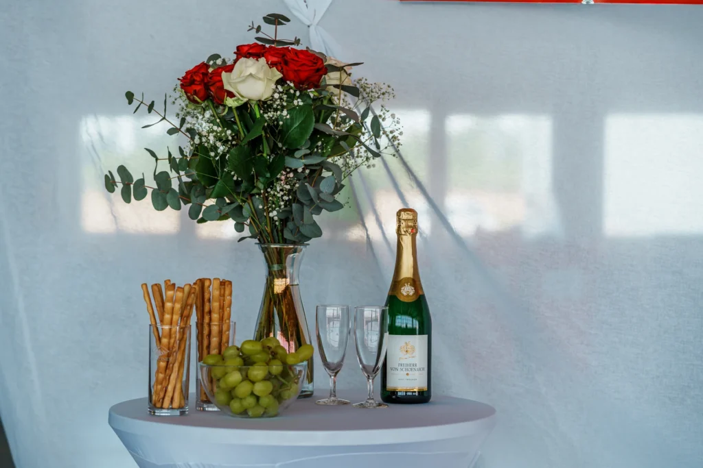 Stehtische vor weißer Leinwand, am Tisch befinden sich Weintrauben und Snacks, sowie eine Flasche Sekt und Gläser.