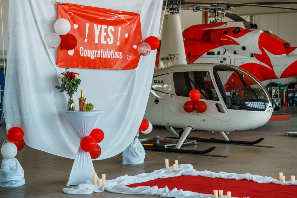 Roter Banner mit der Aufschrift "Yes Congratulation", aufgestellt in einem Hangar. Weißer Hubschrauber mit roten Ballons im Hintergrund.