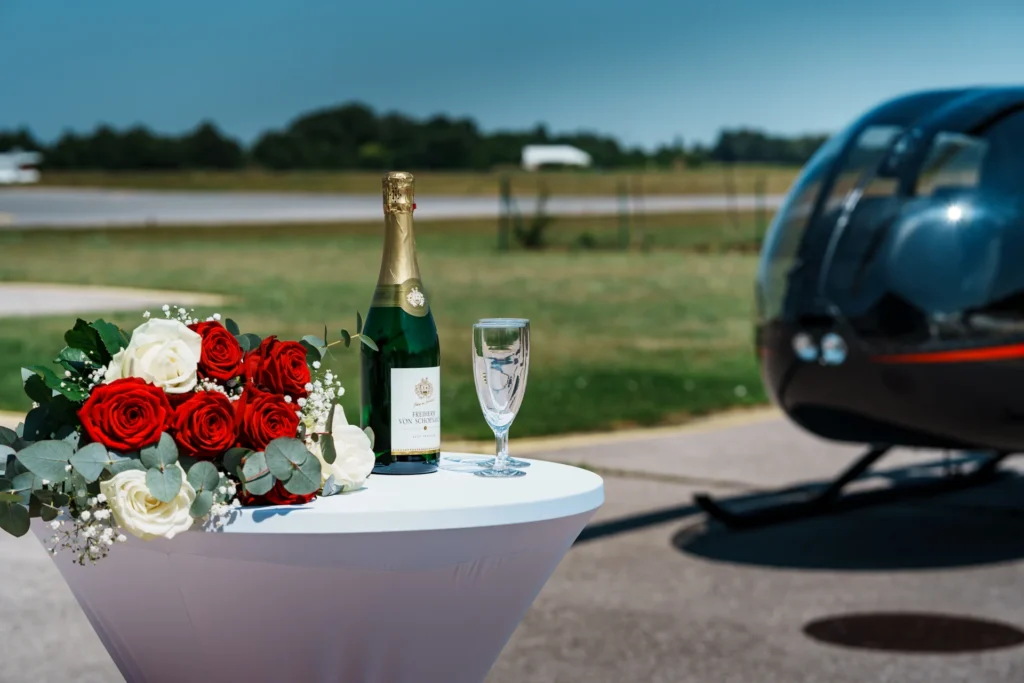 Stehtisch mit Blumenstrauß, Sekt und Sektgläser, im Hintergrund schwarzer Hubschrauber.