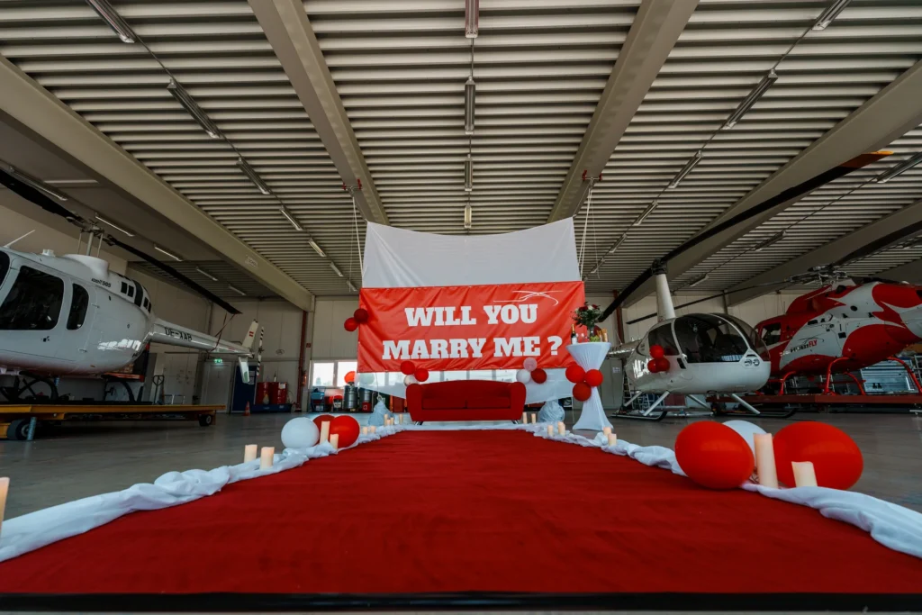 Aufbau in einem Hubschrauber Hangar. Roter Teppich vor einem weißen Vorhang. Rote Couch und ein Stehtisch mit Getränke und Blumen.