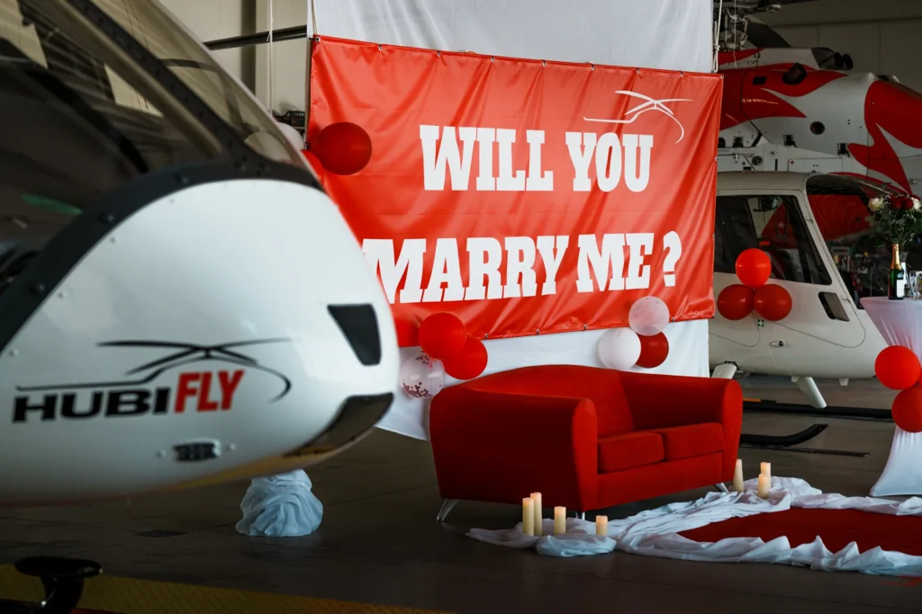 Roter Banner mit der Aufschrift "Will you marry me?", afgestellt in einem Hangar. Weißter Front eines Hubschraubers mit Hubifly-Logo im Vordergrund.