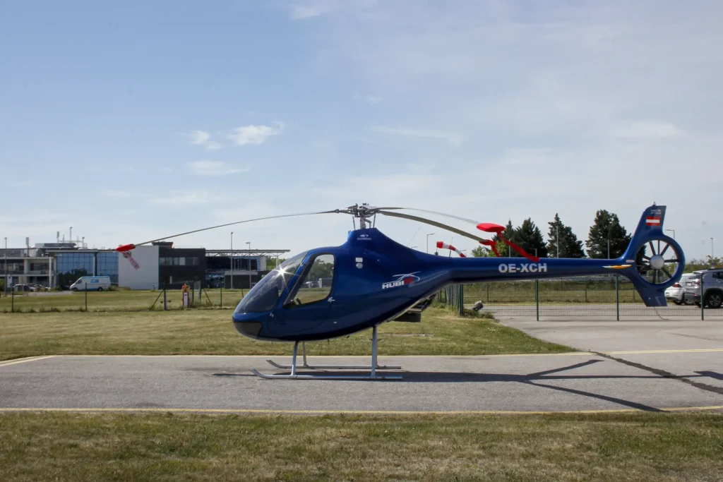 Blue light helicopter with registration OE-XCH parked on a tarmac at an airfield, with buildings and trees in the background.