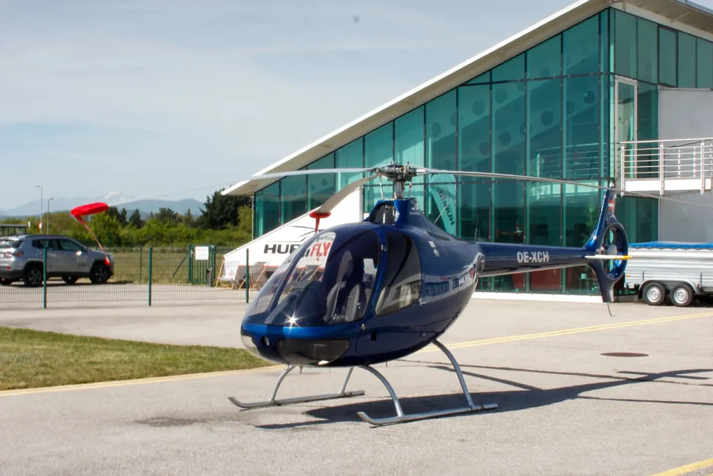 Blue helicopter with skid landing gear sits on a paved apron beside a glass-walled terminal building; red windsock nearby and parked cars in the background.