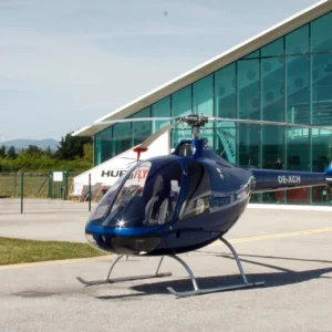 Blue helicopter with skid landing gear sits on a paved apron beside a glass-walled terminal building; red windsock nearby and parked cars in the background.