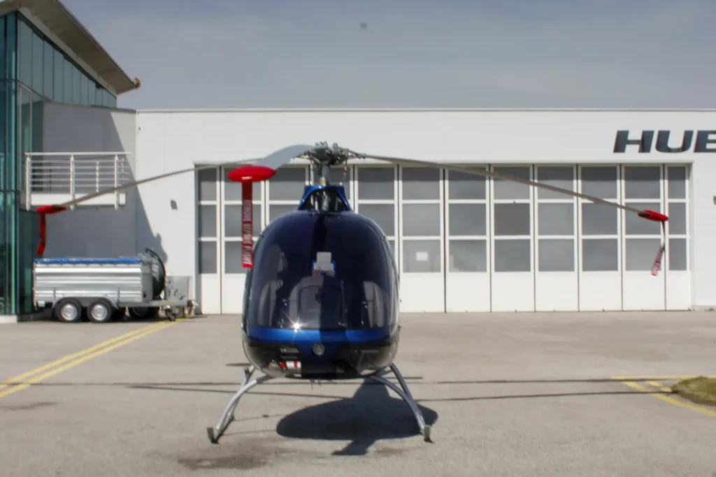 Front view of a blue helicopter with its rotor blades extended, parked on a tarmac in front of a white hangar with glass walls