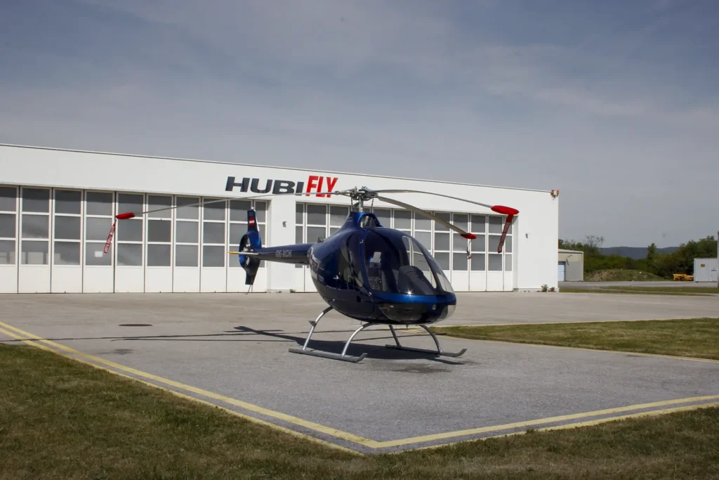 Blue helicopter parked on apron in front of a white hangar with HUBI FLY sign.
