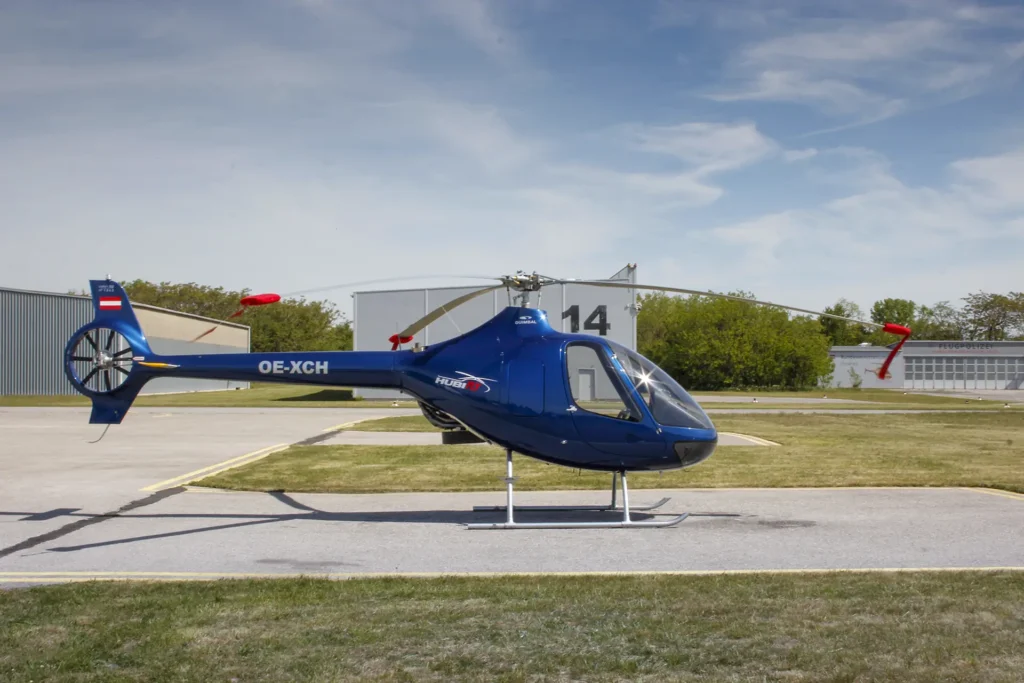 Blue helicopter on a tarmac with its rotor blades at rest, registration OE-XCH visible on the fuselage, and hangars in the background.