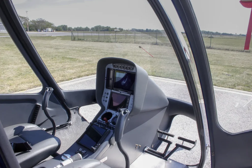 Interior view of a light helicopter cockpit with dual screens, control sticks, and outdoor airfield visible through the open door