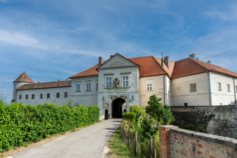 Historic building with red-tiled roof and central arched entrance along a path bordered by green hedges under a blue sky.