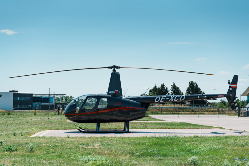 Black and red helicopter parked on a helipad outdoors, registration OE-XCO visible under a clear blue sky.