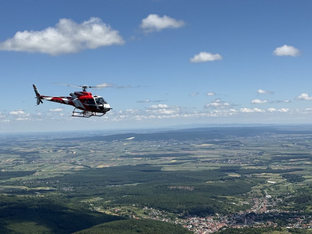 Red helicopter hovering above a flat rural landscape with fields and a blue sky.